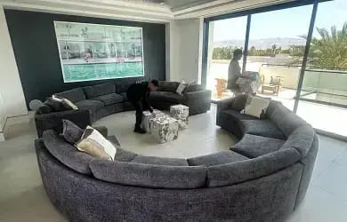 Professional movers arranging a large, semi-circular sectional sofa and ottomans in a modern living room in Palm Desert